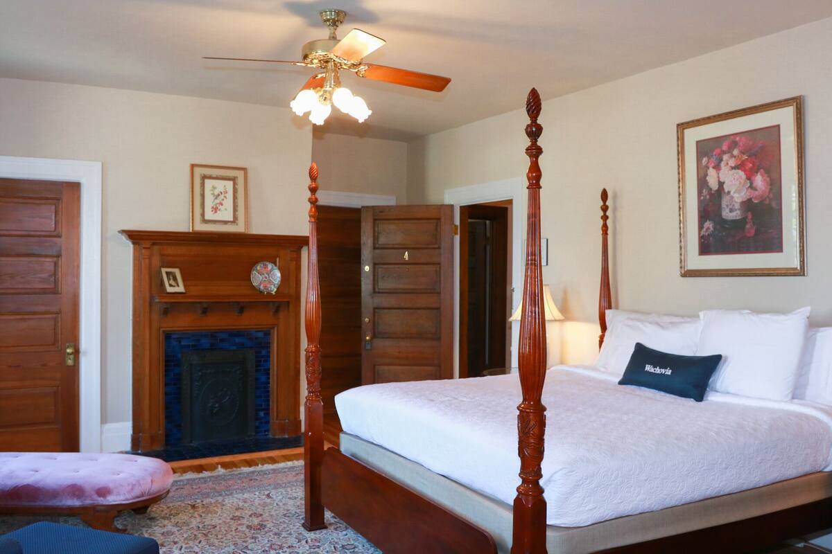 A four-poster bed with a white bedspread, a wooden headboard, and a single decorative pillow.
