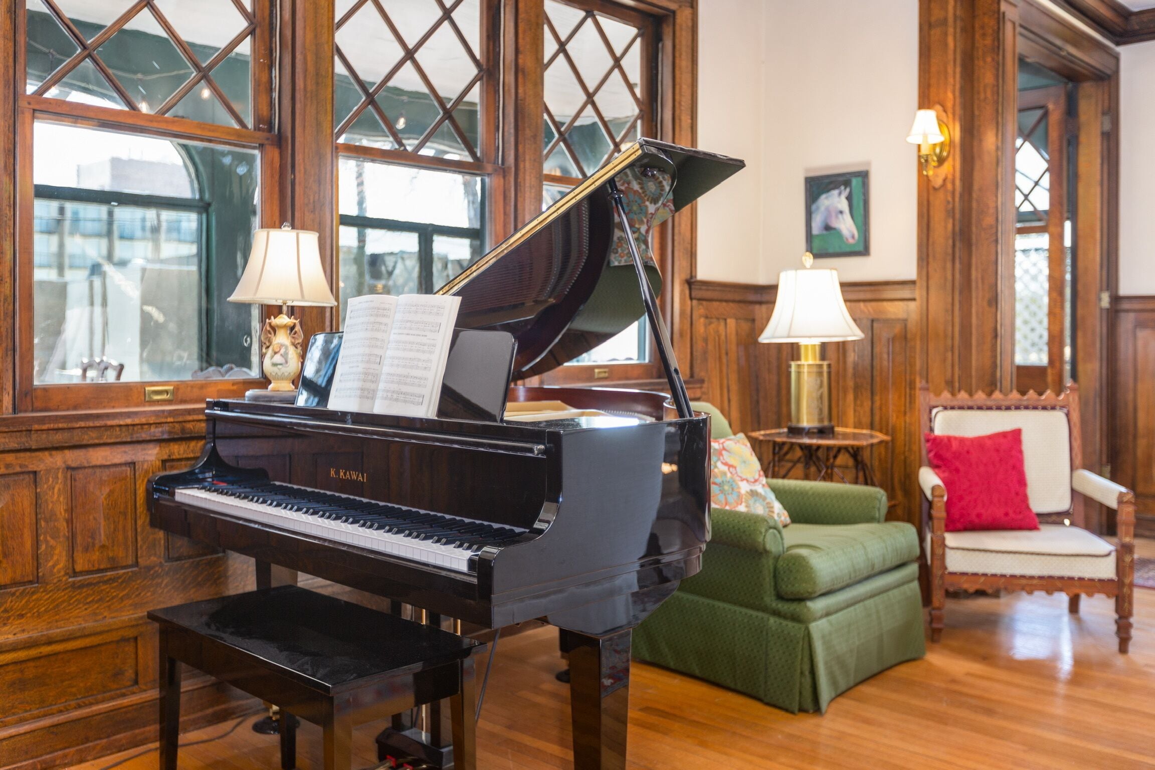 A grand piano in a room with wooden paneled walls, a green armchair, a white chair, and a small table with a lamp.