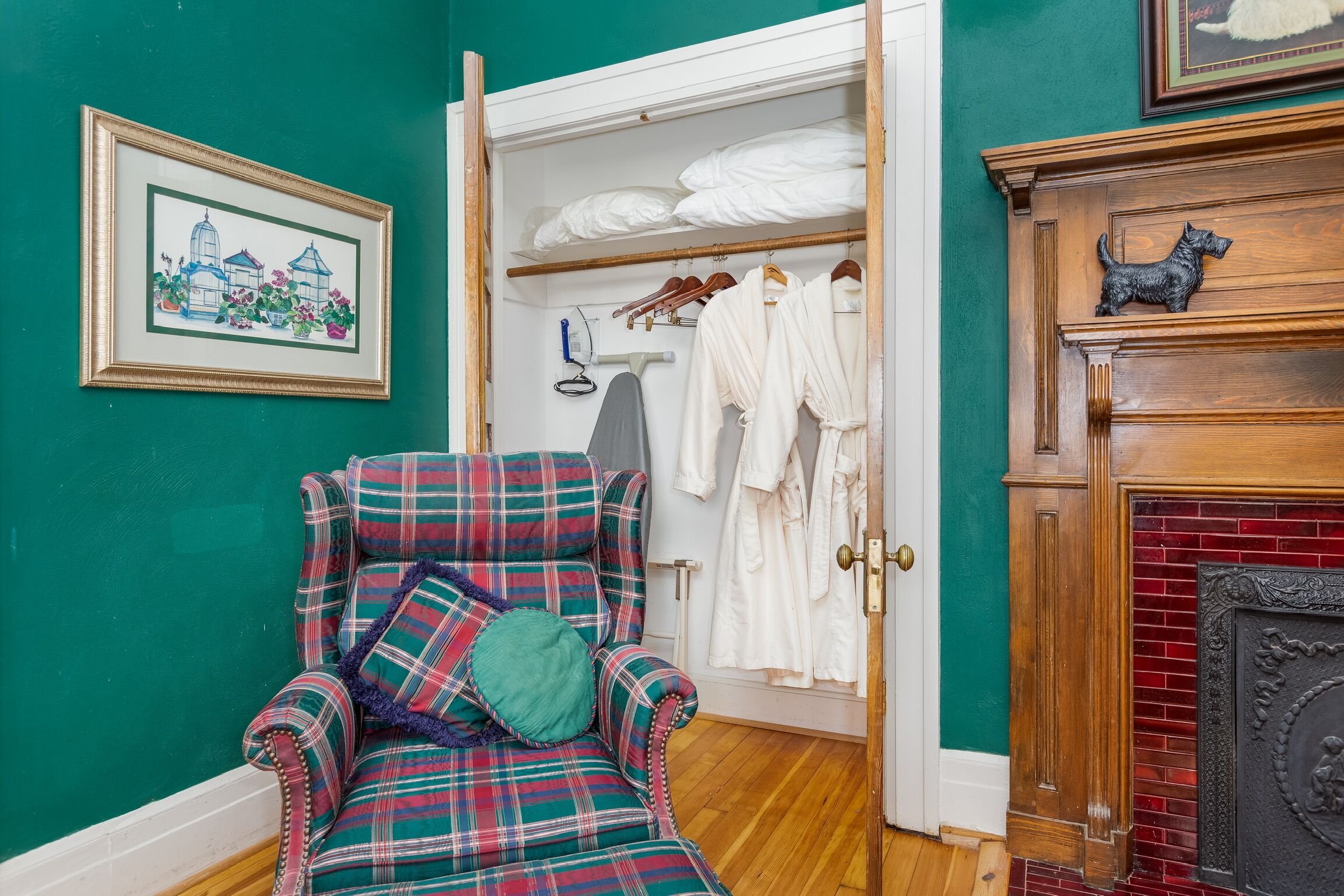 A plaid armchair with a cushion, a wooden cabinet with a dog sculpture, and a white door leading to a closet with hanging clothes and a framed picture on the wall.