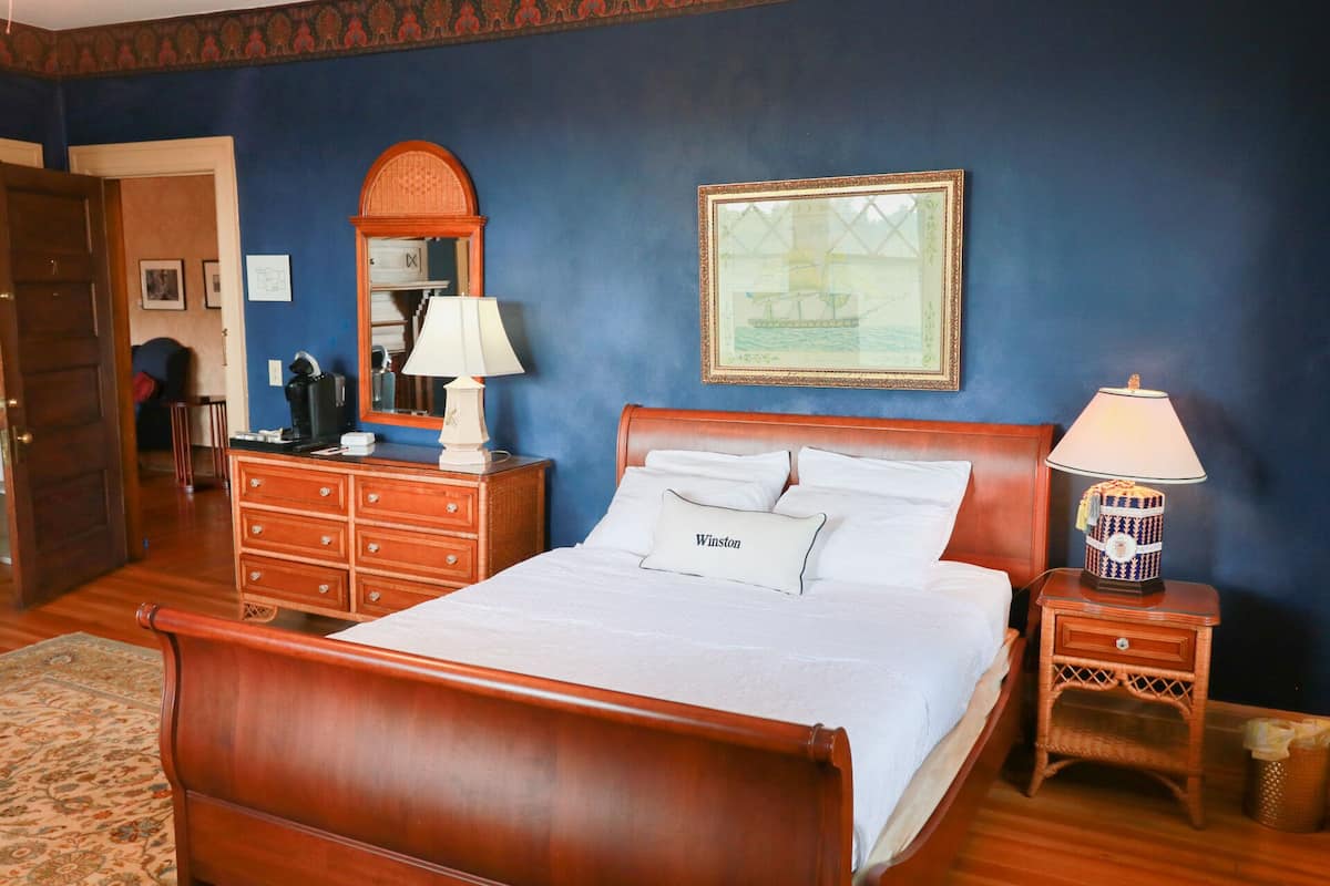 A hotel room with a wooden bed, a nightstand with a lamp, a dresser, a mirror, and a framed picture on the wall.
