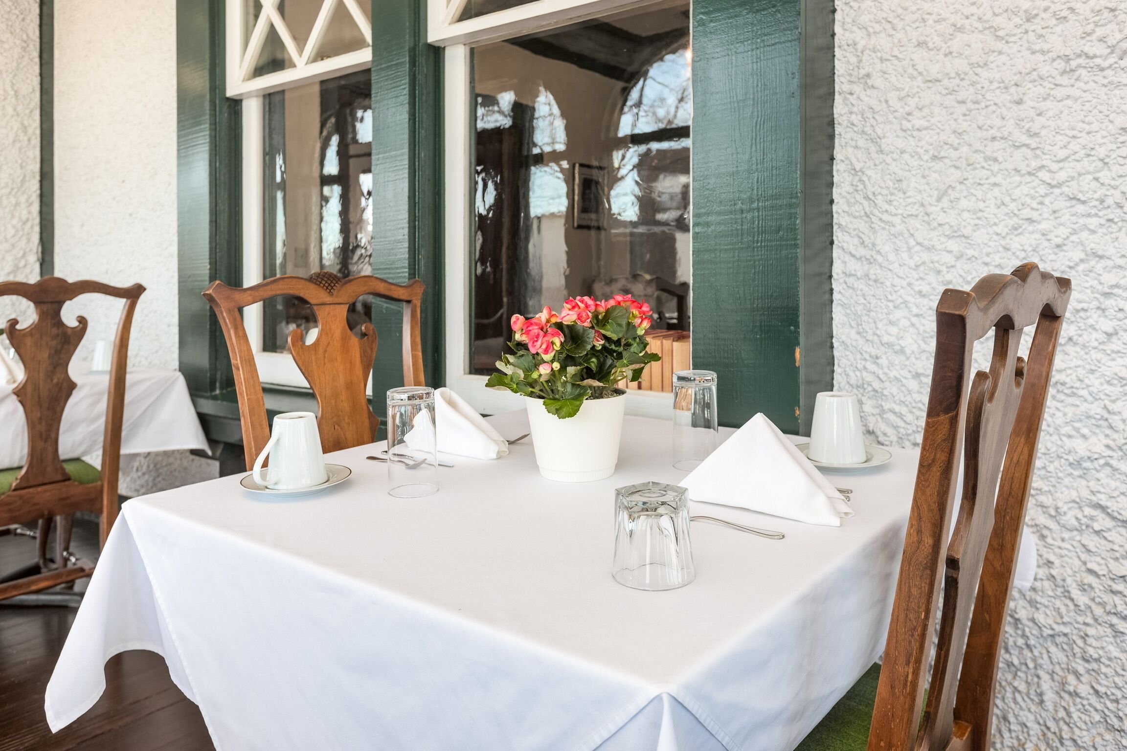 A table set for two with a white tablecloth, a white vase with pink flowers, and wooden chairs.