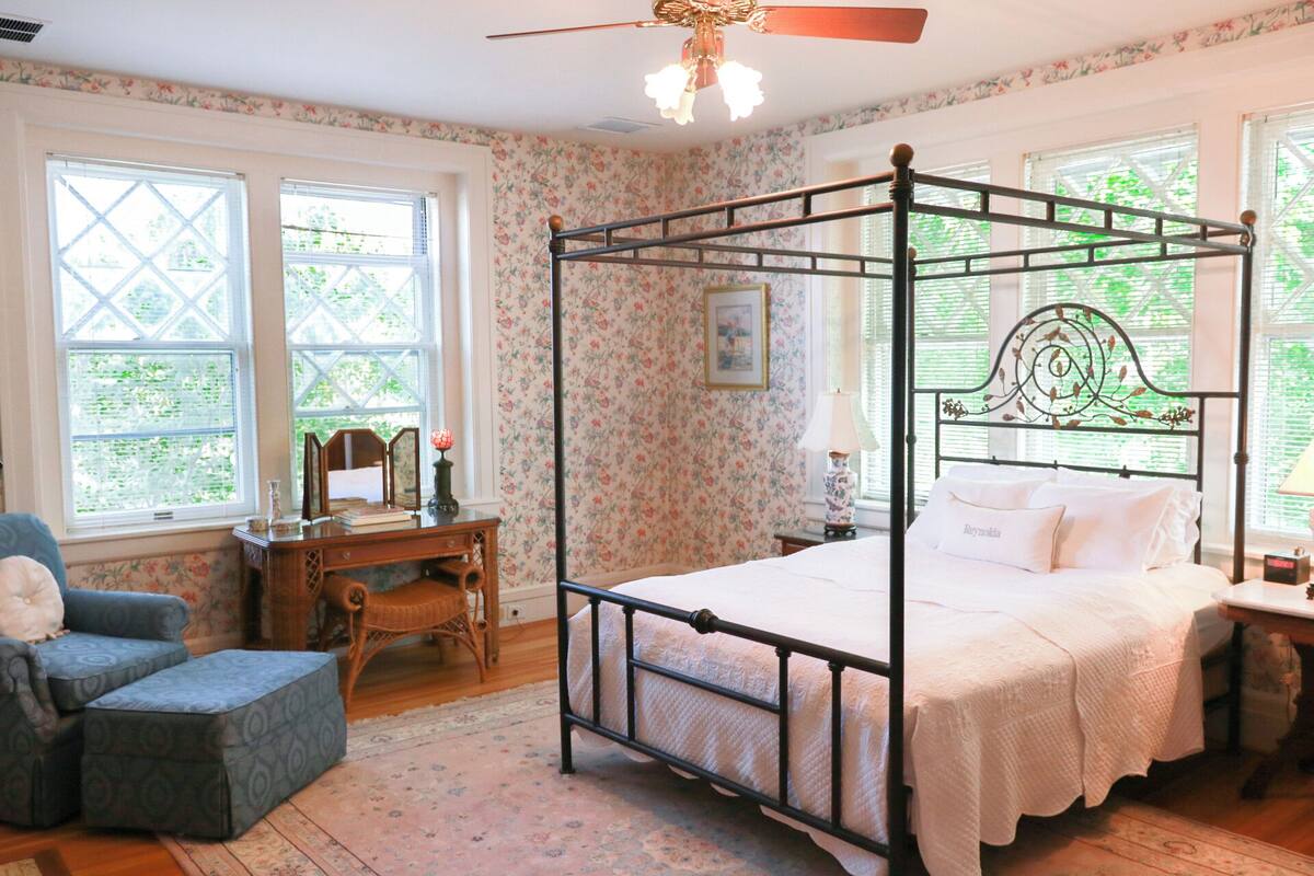 A four-poster bed with a white bedspread, a wooden desk with a chair, and a window with a floral wallpaper pattern.