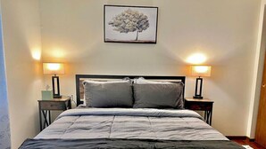 Un letto ben rifatto con copriletto grigio e nero a righe, due lampade da comodino e un quadro con un albero appeso al muro.