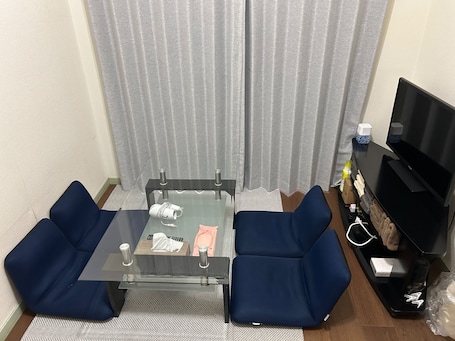 A small living area with a glass-top table, two blue chairs, a TV, and a shelf with various items.