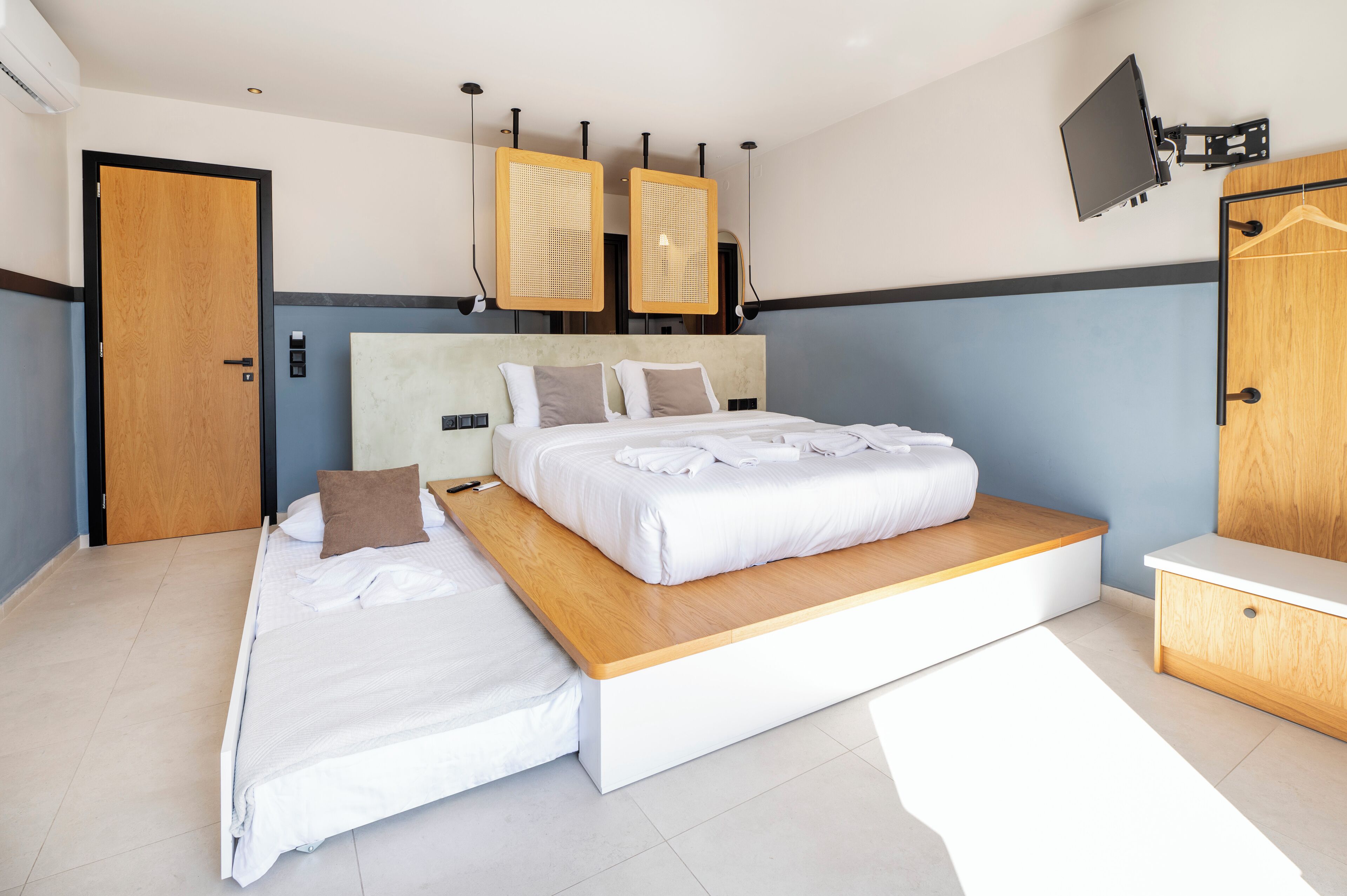 A modern hotel room with a large bed, a wooden headboard, a flat-screen TV mounted on the wall, and a wooden wardrobe.