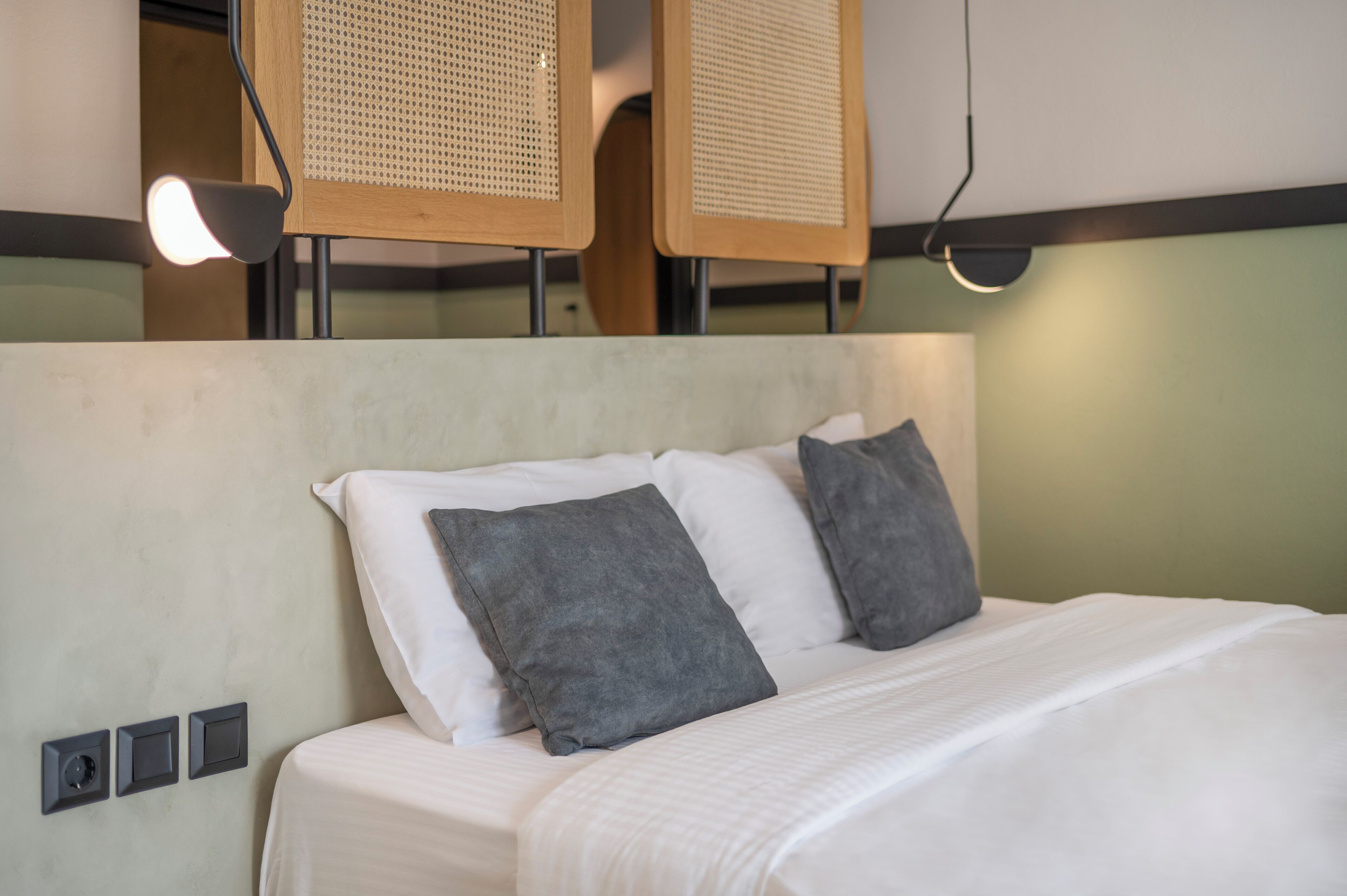 A neatly made bed with white and gray bedding, a headboard with a woven pattern, and a wall-mounted light fixture.