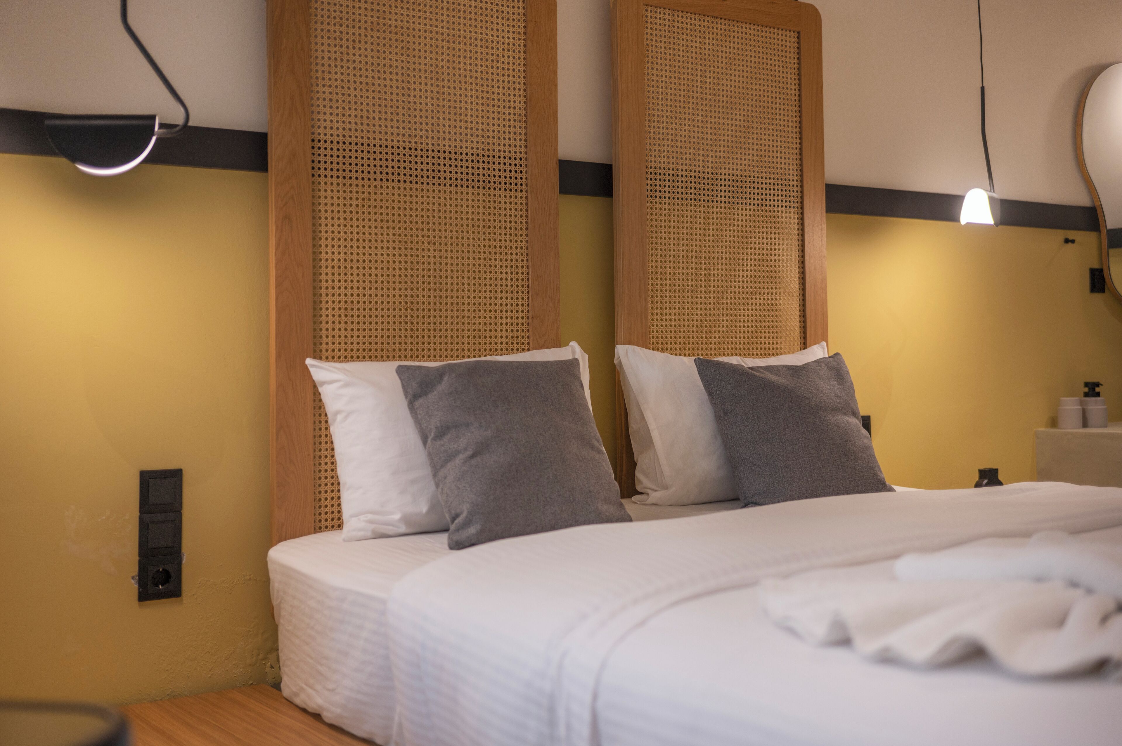 A neatly made bed with white linens and two grey pillows, a wooden headboard, a bedside table with a lamp, and a wall-mounted light fixture.
