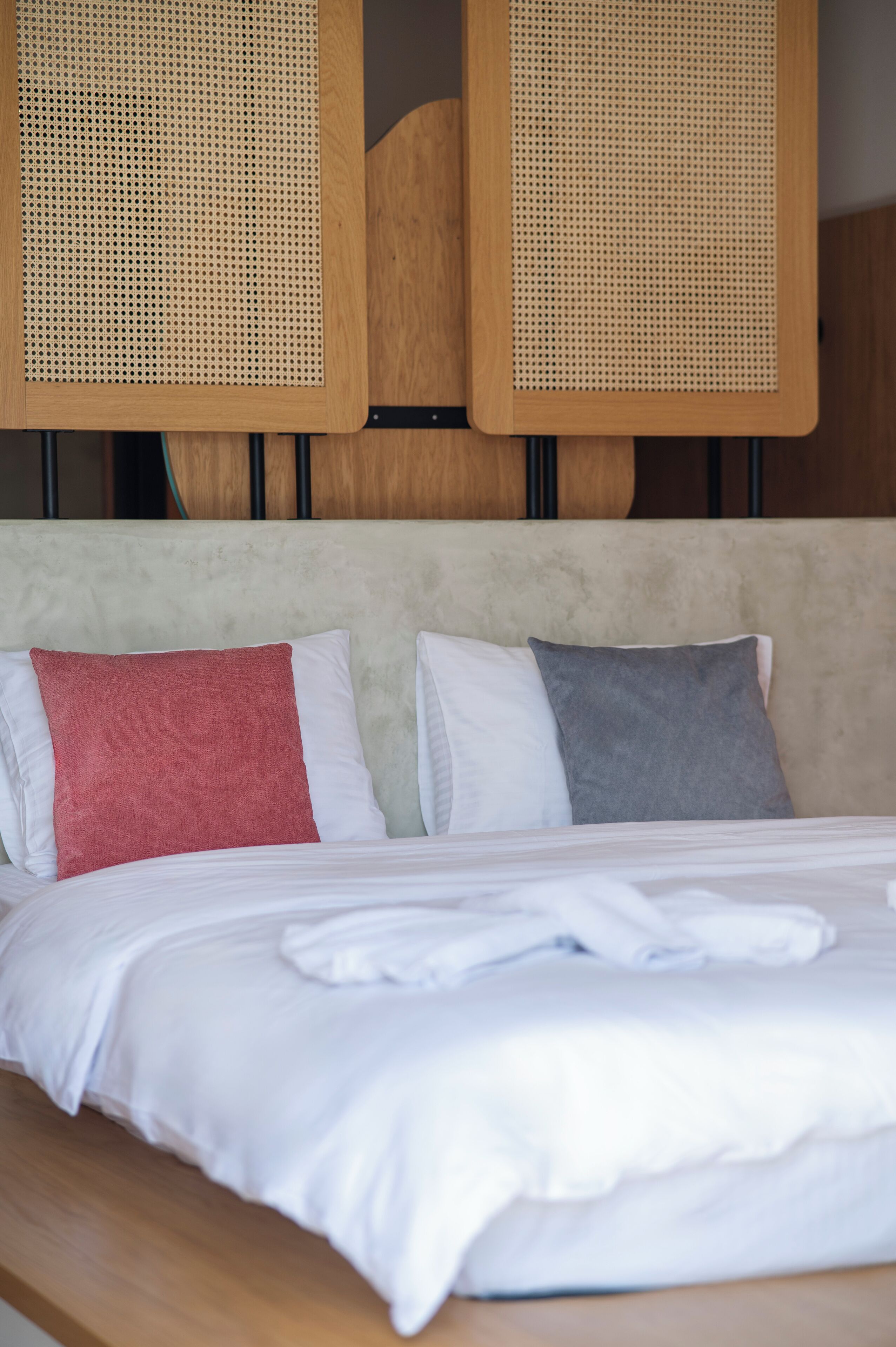 Una cama bien hecha con ropa blanca y dos almohadas, una roja y otra gris, en una habitación con cabecera de madera y dos cabeceras tejidas sobre la cama.