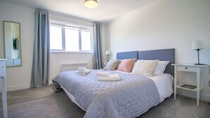 Ein ordentlich eingerichtetes Schlafzimmer mit einem großen Bett, einem Nachttisch, einer Lampe und einem Fenster mit Vorhängen.