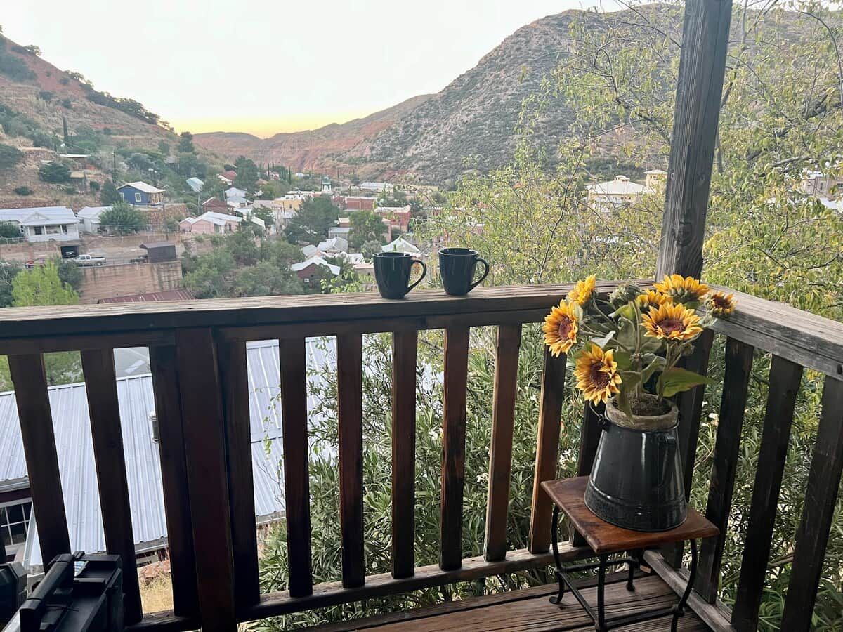 Un balcón con barandilla de madera, una planta en maceta con girasoles y dos tazas negras.