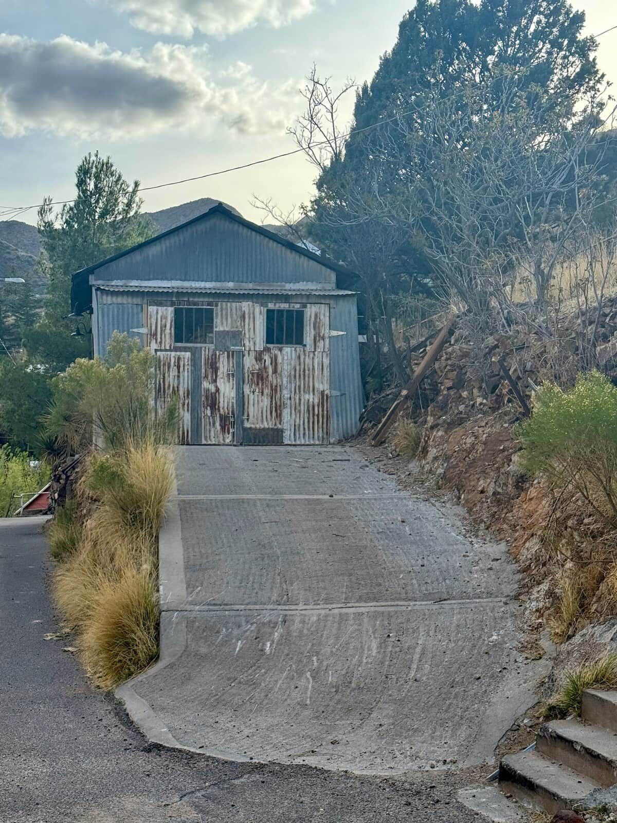 A weathered building with a corrugated metal roof, a concrete driveway leading to a gated entrance, and a hillside in the background.