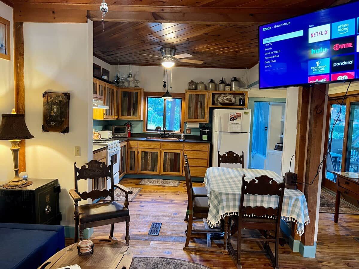 Un salon chaleureux avec un plafond en bois, un téléviseur fixé au mur, une table à manger avec des chaises et, au fond, un coin cuisine.