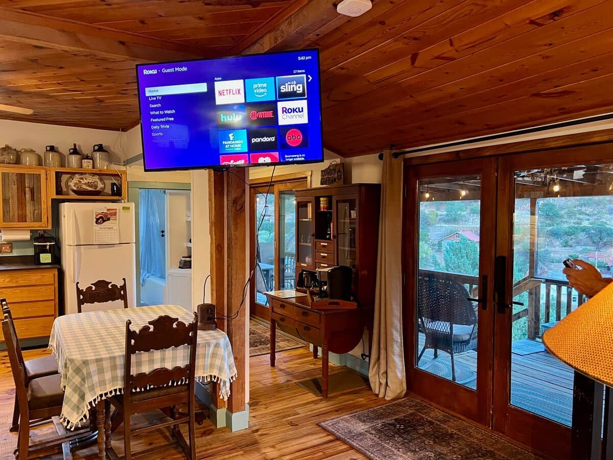 A wooden interior with a television displaying a guest mode menu, a dining area with a table and chairs, and a view of the outdoors through a glass door.