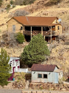 A two-story wooden house with a balcony, surrounded by dry vegetation and a large tree. - Top of the Stairs Old Bisbee|Park & Walk to Main Street (Bisbee)