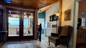 A cozy living room with a wooden ceiling, a comfortable armchair, a small table with a remote control, and a view of a balcony with a wooden railing. - 3-bedroom cabin in Old Bisbee (Bisbee)