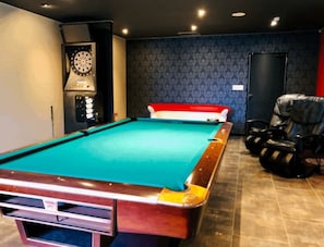 A pool table with a dartboard and a red and white sofa in a room with patterned wallpaper and a door.