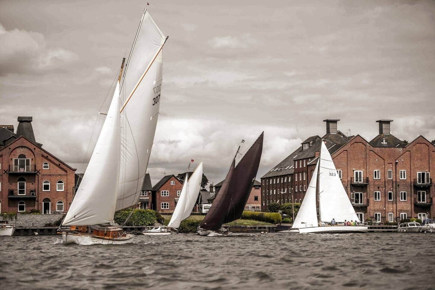 A sailboat with a white sail and a dark sail in a harbor with brick buildings in the background.