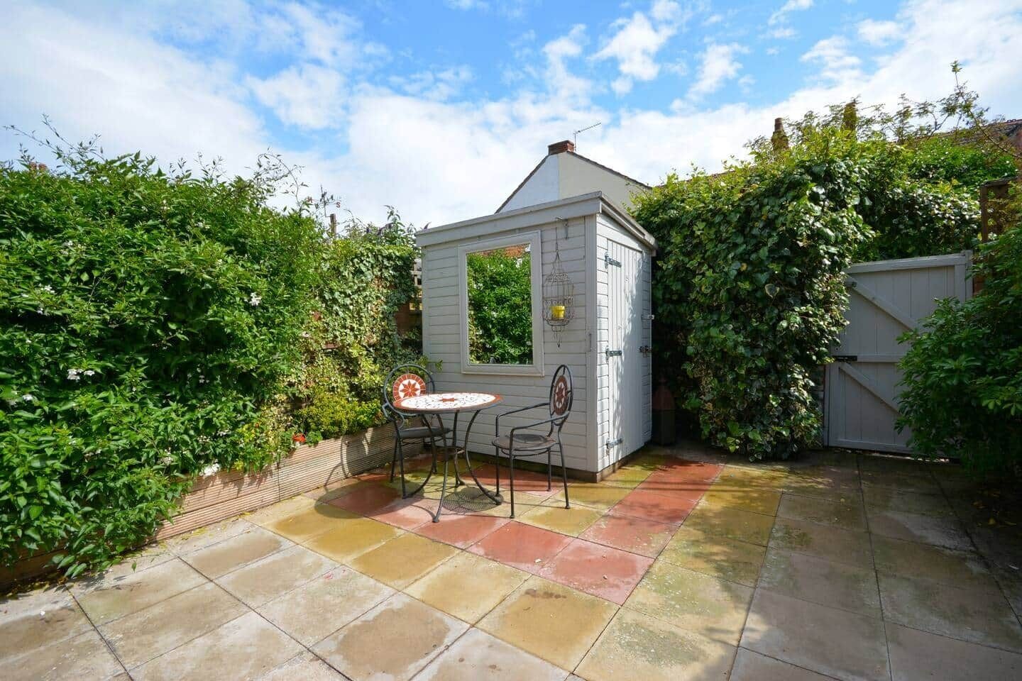 A small, white wooden structure with a window, surrounded by lush greenery and a tiled patio area with two chairs.