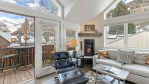 Ein geräumiges Wohnzimmer mit Kamin, einer Couch, einem Couchtisch und einem Sessel. Eine Terrasse mit Gartenmöbeln und Blick auf Berge.