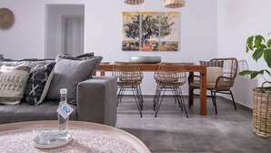 A modern living room with a grey sofa, wicker chairs, a wooden dining table, and a large painting on the wall. - Infinite Blue Apartments (Malevizi)