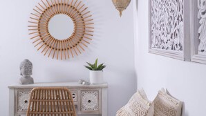 A room with a white wall, a round wooden wall art, a white cabinet with drawers, a wicker chair, a macrame pillow, and a hanging light fixture. - Infinite Blue Apartments (Malevizi)