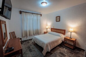 A bedroom with a bed, two bedside tables with lamps, a wooden desk, a chair, a mirror, and a window with curtains.