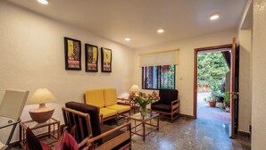 A hotel lobby with a yellow sofa, wooden chairs, a glass coffee table, and a lamp. There are framed pictures on the wall and a door leading to an outdoor area with plants. - Bungalows San Felipe (Oaxaca)
