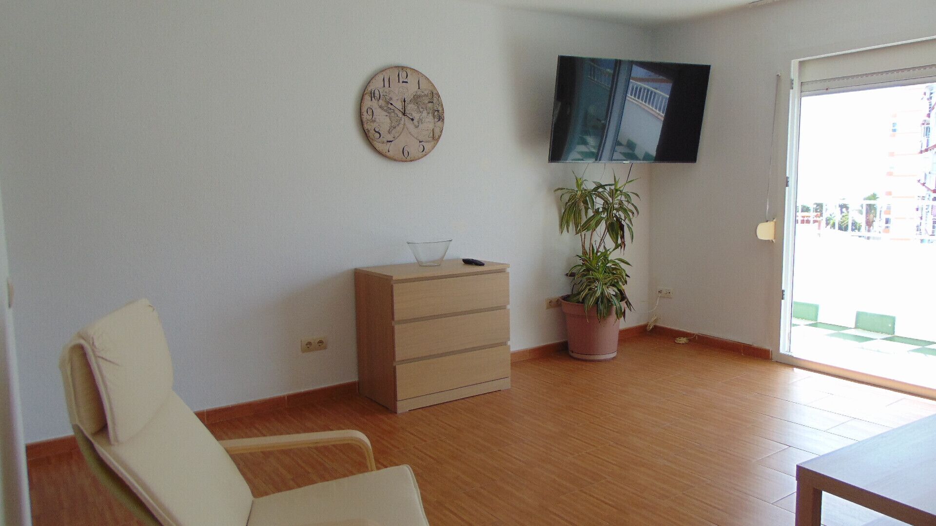 Ein Zimmer mit Holzboden, einem beigefarbenen Sofa, einem Holzschrank, einem Blumentopf, einer Wanduhr und einem an der Wand befestigten Fernseher.