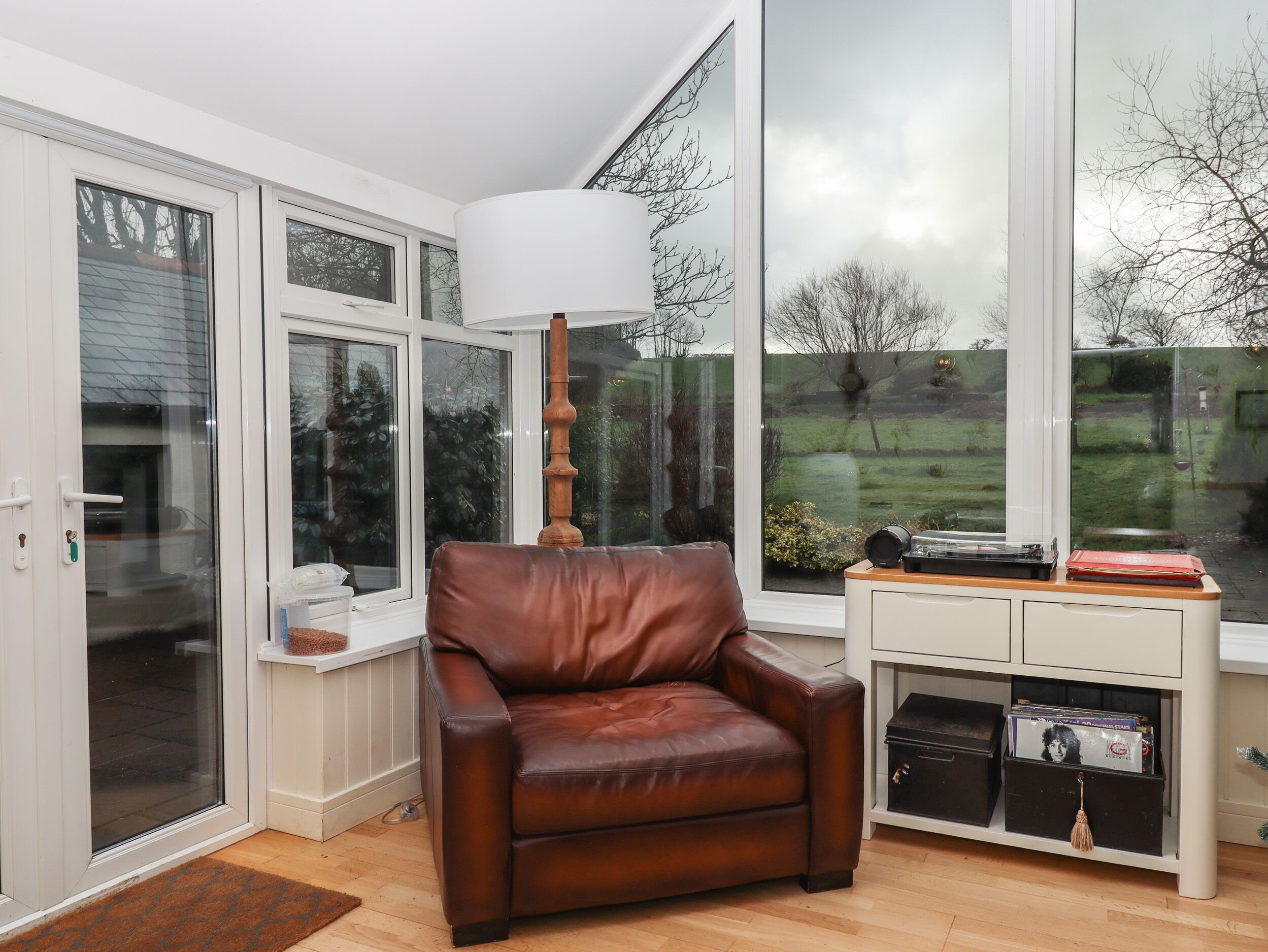 A room with a brown leather chair, a white cabinet with records, and large windows offering a view of a green landscape.