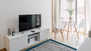 Een moderne woonkamer met een flatscreen televisie op een wit kastje, een glazen eettafel met witte stoelen en een groot raam met dunne gordijnen.