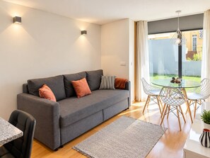 A modern living room with a grey sofa, a glass dining table, and chairs. There are two wall-mounted lights, a large window with curtains, and a view of the outside.