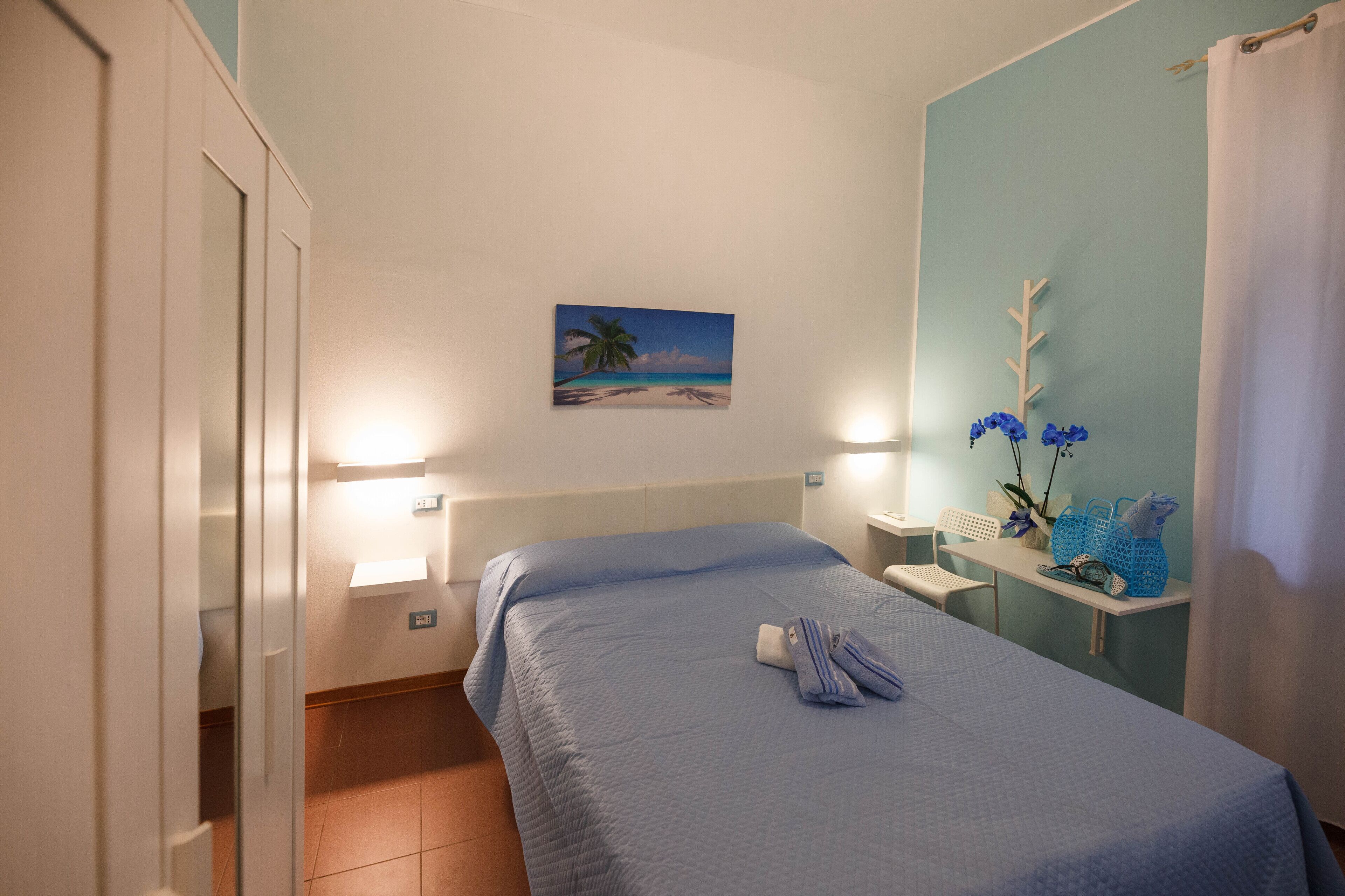 Ein Hotelzimmer mit einem Bett, einem Strandbild, einem kleinen Tisch mit Blumen und einem Sessel.