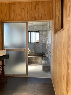 Ein Zimmer mit Holzwand, einer Schiebetür zu einem Badezimmer mit Marmorwand, einem vergitterten Fenster und einem Waschbecken.
