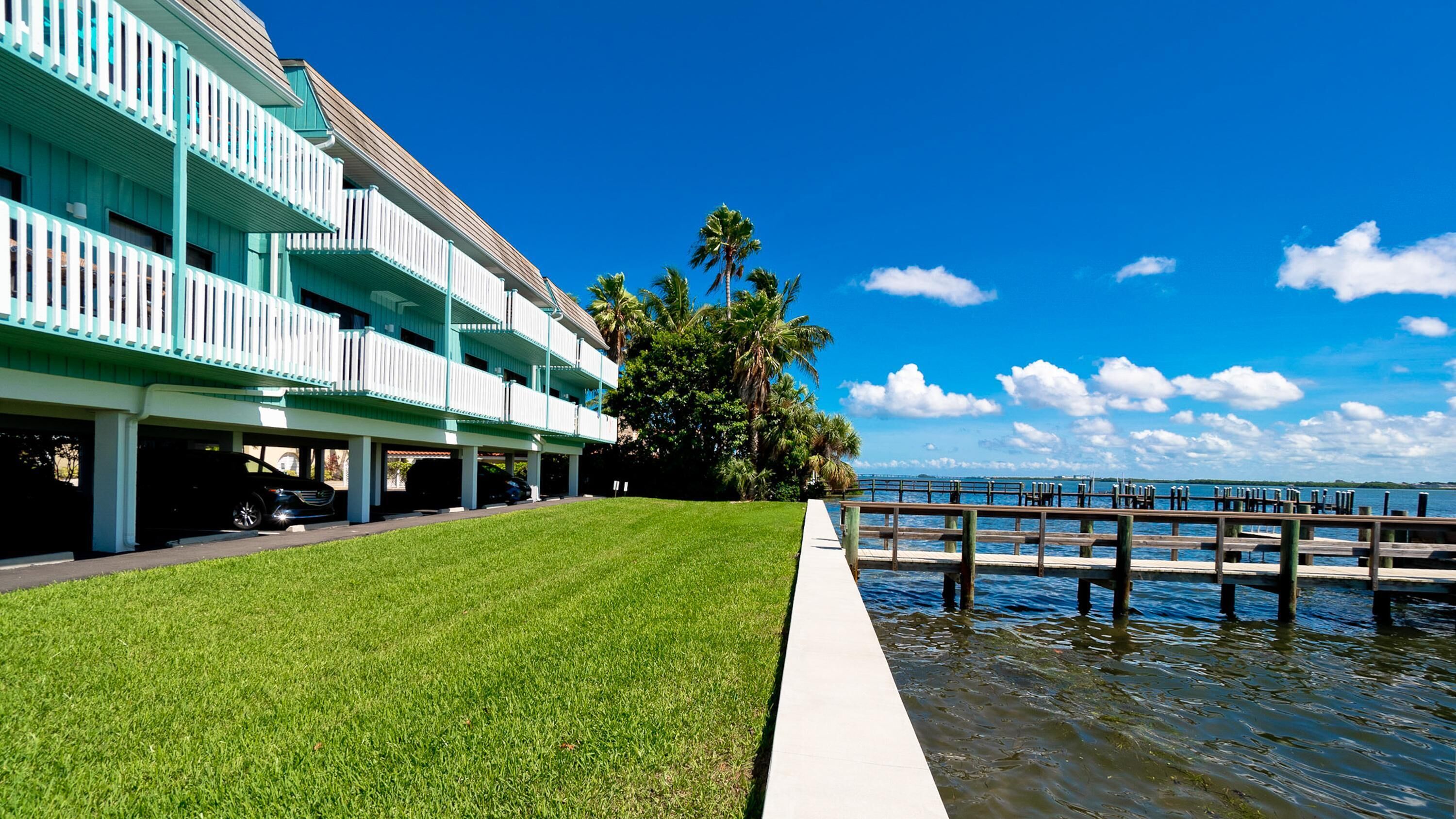 A hotel with a grassy lawn, a wooden dock extending into the water, and a clear blue sky.