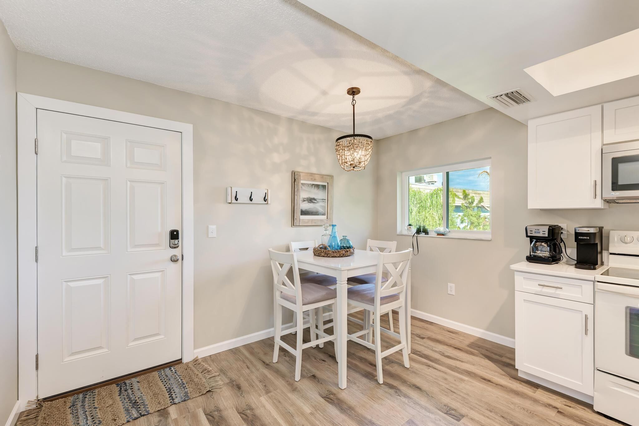 A kitchen with white cabinets, a dining table with chairs, a chandelier, and a window with a view of greenery.