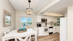 A modern kitchen with white cabinetry, a dining table with blue glassware, and a view of the outdoors. - Anna Maria Island Beach Palms 8B (Bradenton Beach)