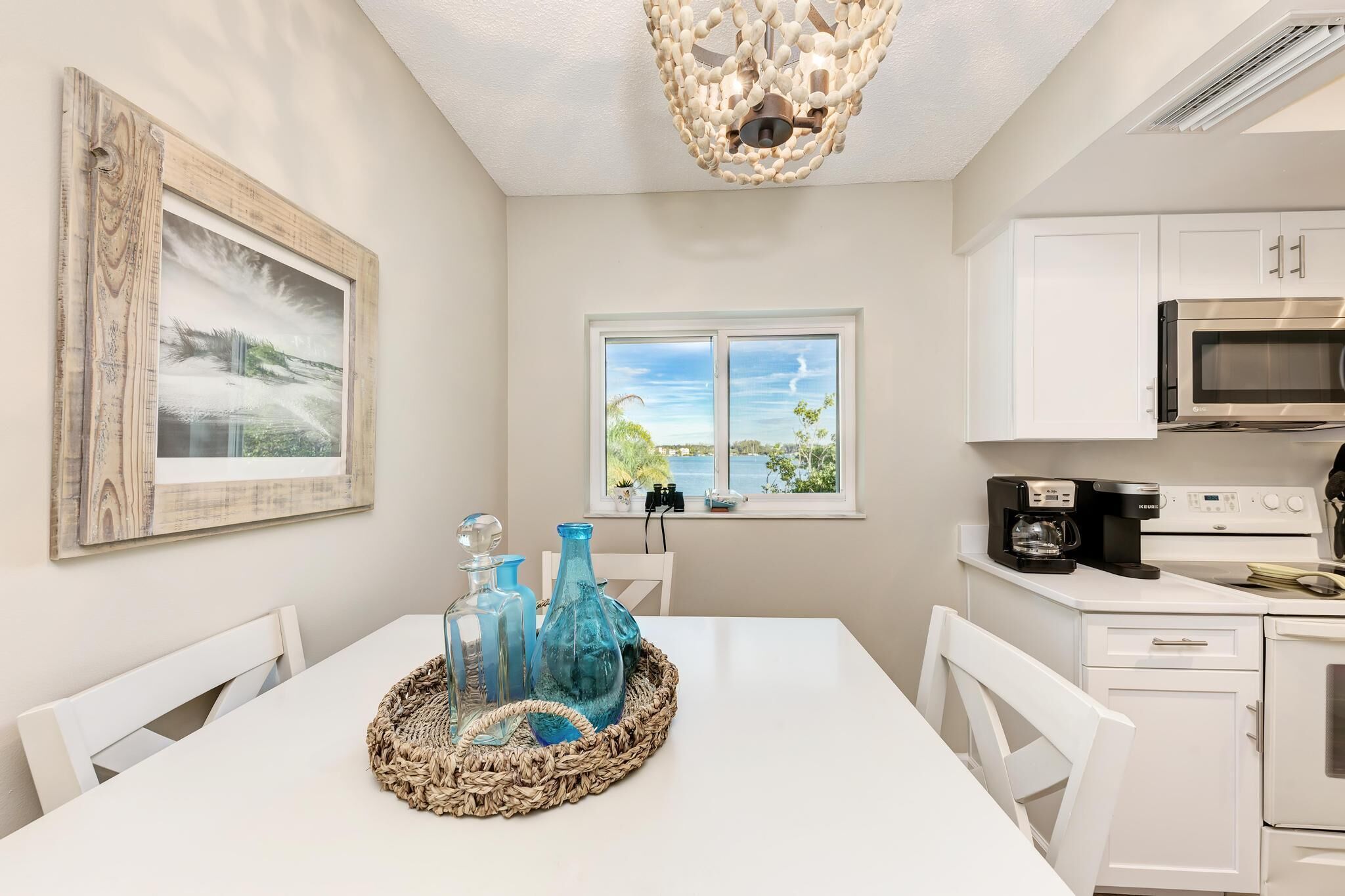 A dining area with a white table, blue glassware, and a woven placemat. There is a framed picture on the wall, a window with a view, and a kitchen with white cabinets and appliances.