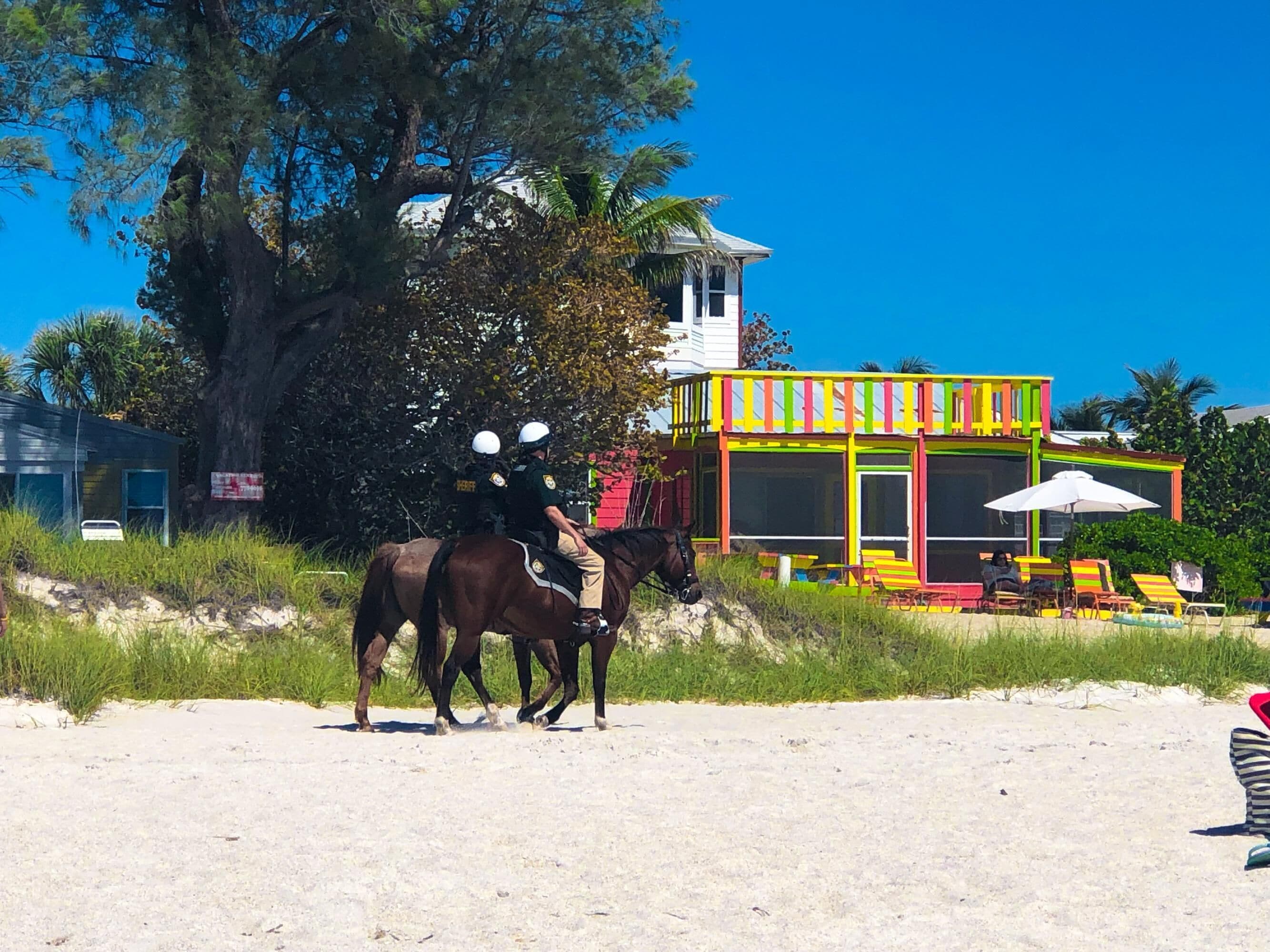 A beach scene with a colorful building, two police officers on horseback, and beach chairs.