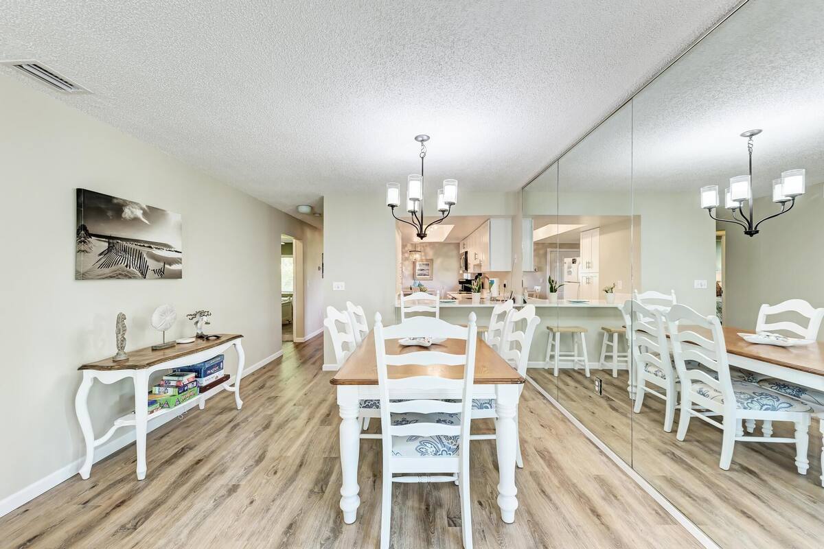 A dining area with a wooden table and white chairs, a chandelier, and a large mirror reflecting the kitchen.