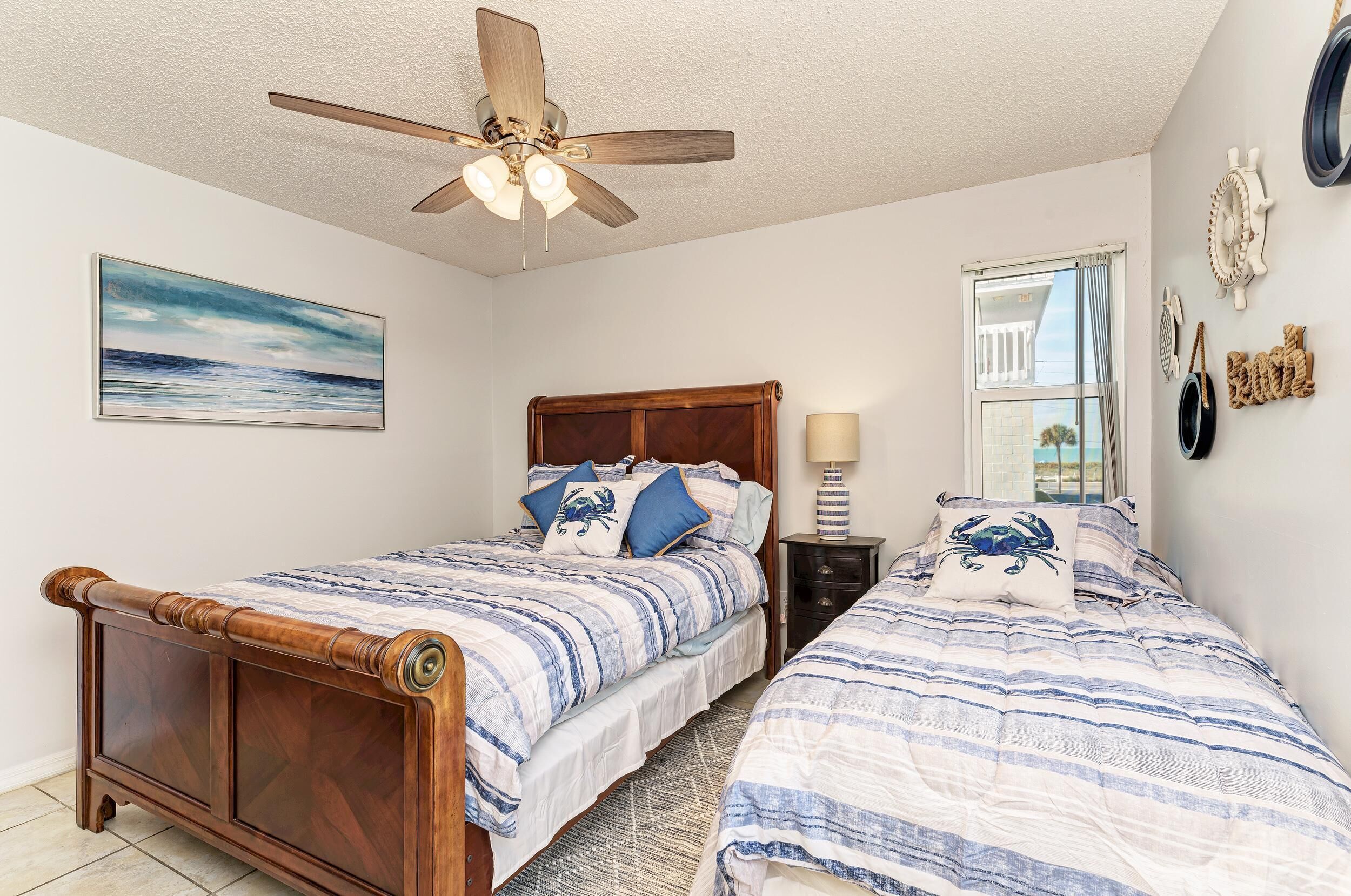 A bedroom with two beds, a ceiling fan, a window with curtains, and nautical-themed decor.