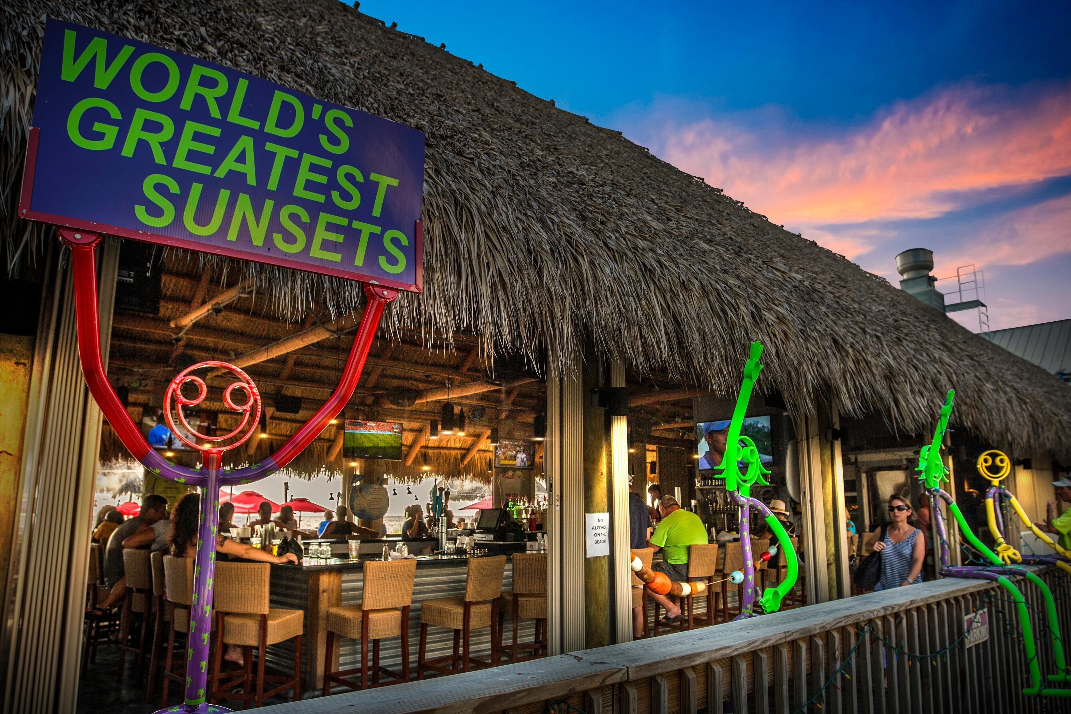 Un bar situé en bord de mer, doté d’un toit de chaume et d  ’une enseigne sur laquelle on peut lire : « LES PLUS BEAUX COUCHERS DE SOLEIL DU MONDE ».