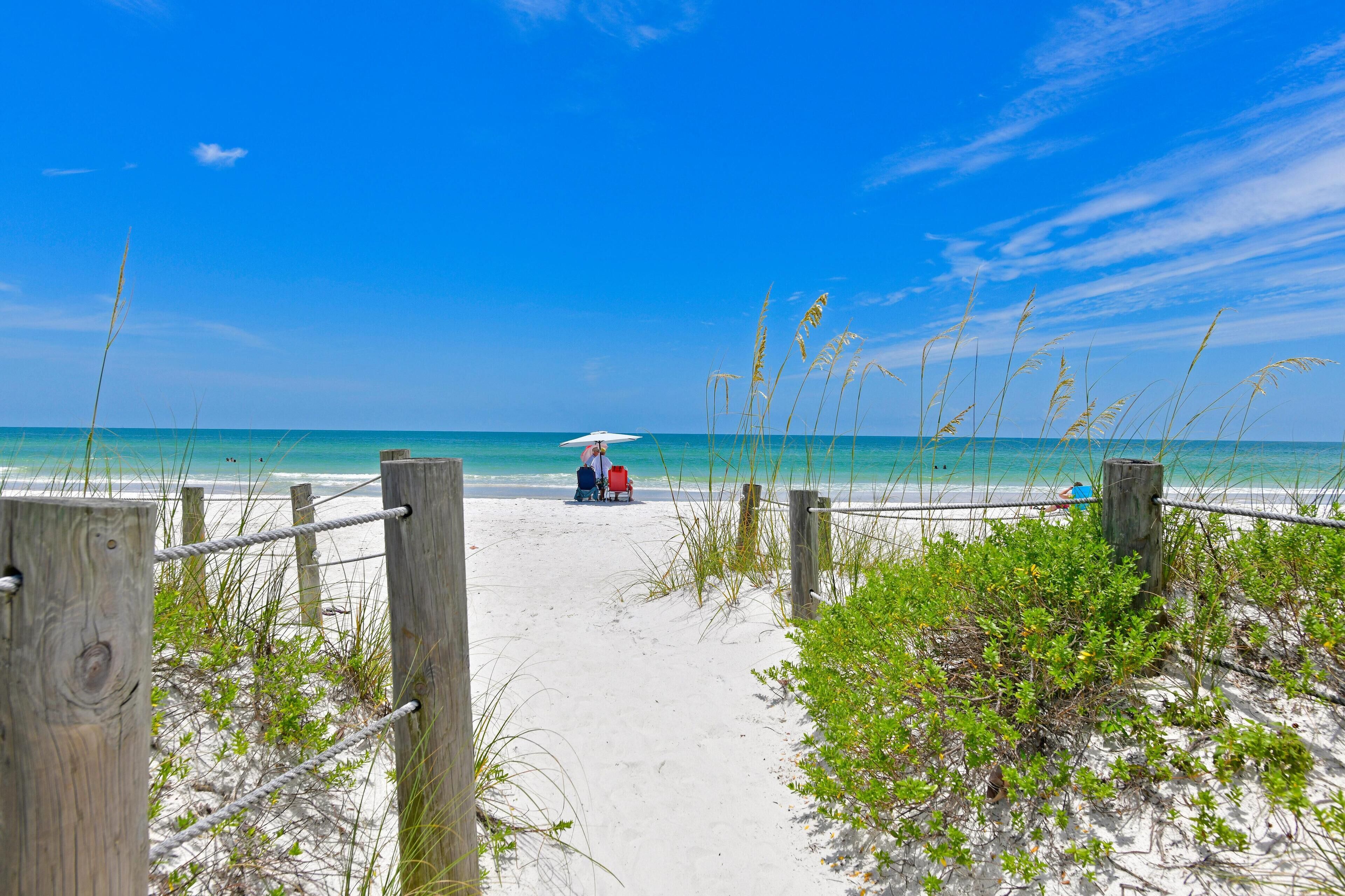 A sandy beach path leading to the ocean with a wooden fence and some vegetation.