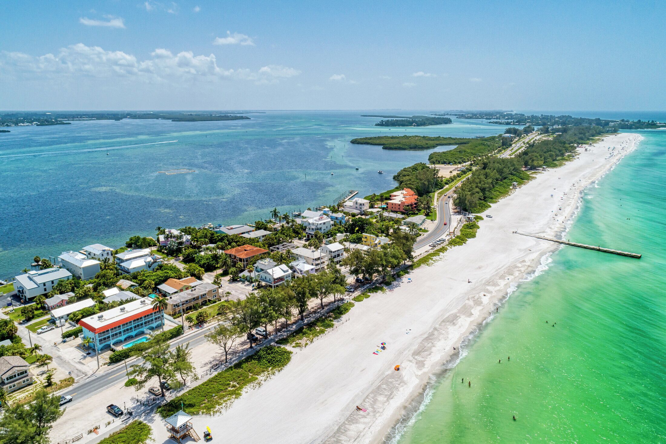 Luchtbeeld van een kustplaatsje met een lang zandstrand, helder turquoise water en een combinatie van woon- en bedrijfsgebouwen.