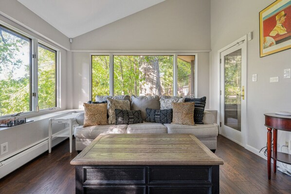 A modern living room with a large window, a sofa with patterned cushions, a wooden coffee table, and a painting on the wall.