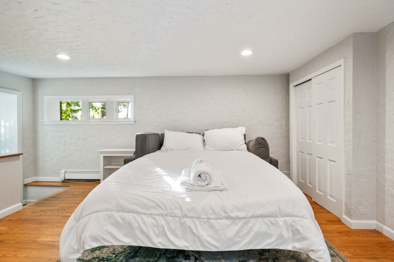 A bedroom with a large bed, a nightstand, a window with a view of trees, and a white door.