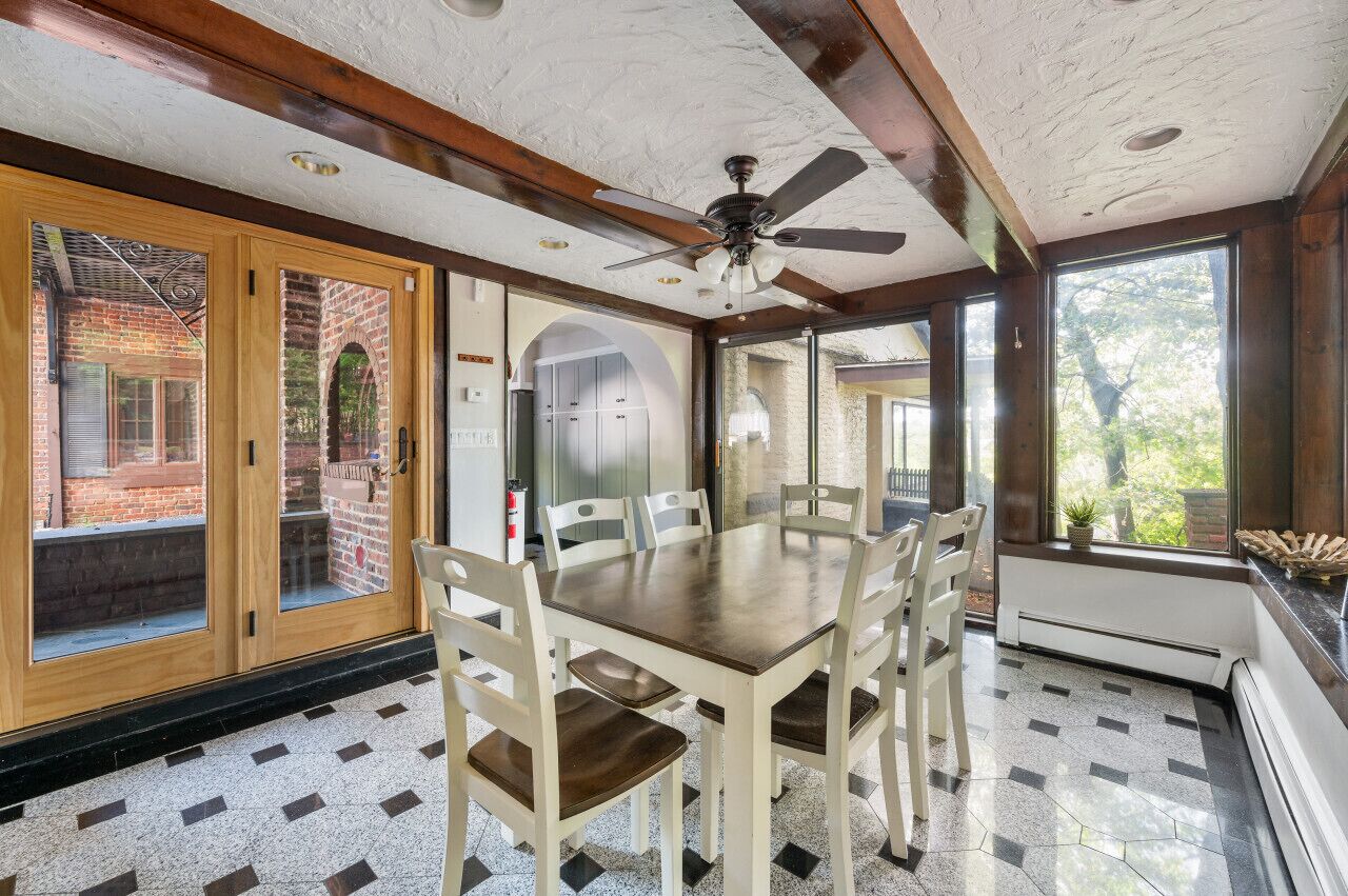 Uma área de refeições com mesa e cadeiras de madeira, um ventilador de teto e janelas amplas com vista para o exterior.