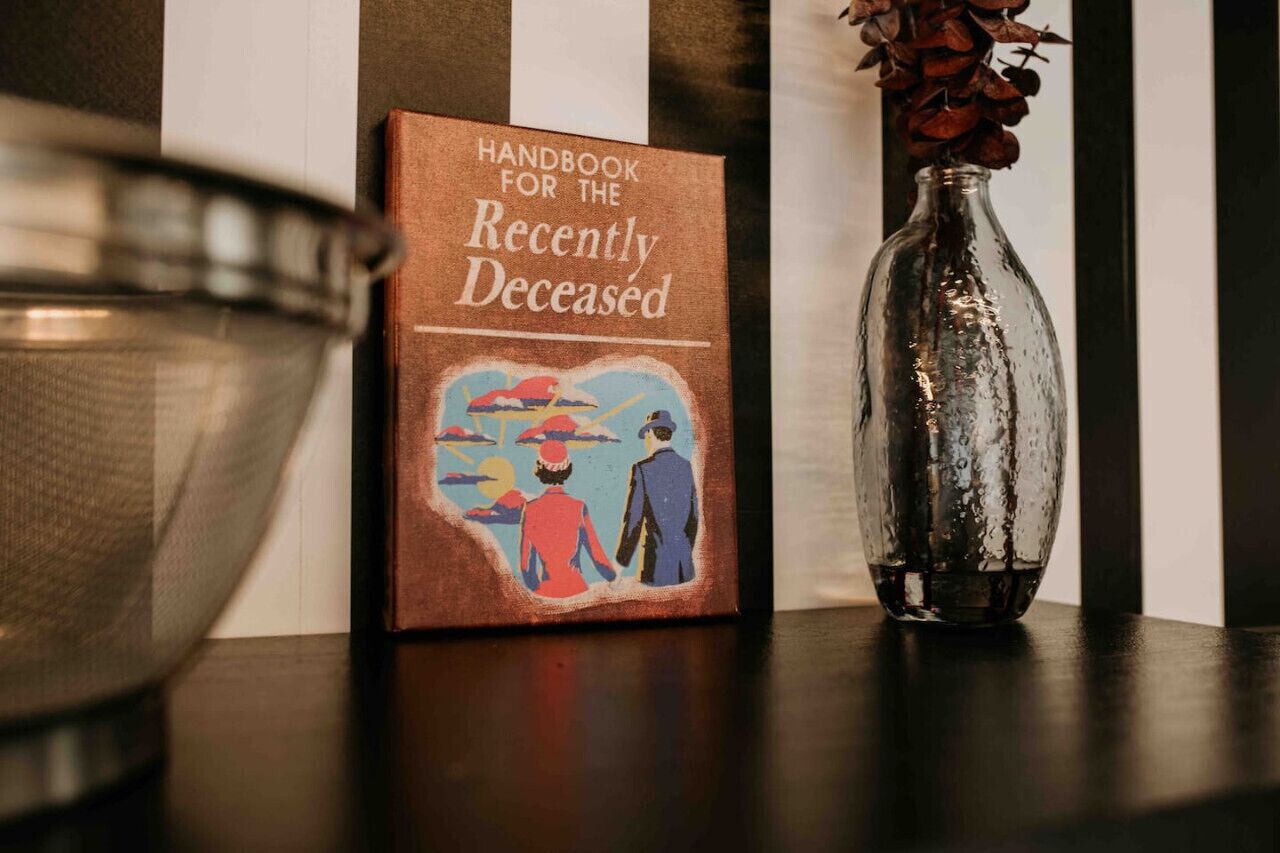 Un libro intitolato 'Handbook for the Recently Deceased' su uno scaffale, accanto a un vaso di vetro con fiori secchi.