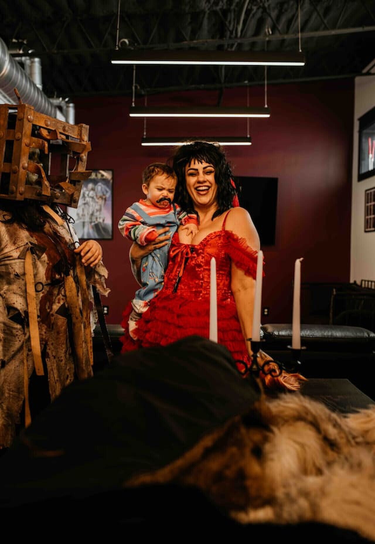 A room with a bed, a wooden structure, and a person in a red dress holding a child.