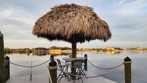 A wooden dock with a thatched umbrella table and chairs, a rope barrier, and calm water in the background.