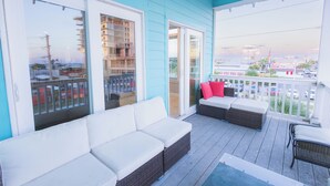 A covered outdoor area with a white sofa and wicker furniture, large sliding glass doors leading to a balcony, and a view of the cityscape.